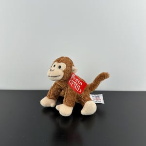 May include: A small brown plush monkey toy with a cream-coloured face, hands, and feet. The monkey features black plastic eyes and a stitched mouth. A red tag attached to the ear reads Gotta Getta GUND, and a white tag on the back reads TELUS GUND.