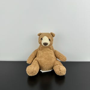Puede incluir: Un oso de peluche sentado. El oso tiene un cuerpo marrón claro, con un hocico y orejas internas más claros. La nariz es de un marrón más oscuro. El oso está sentado sobre una superficie negra, con una etiqueta blanca visible.
