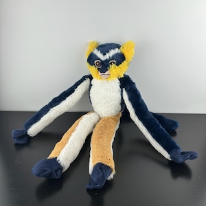 May include: A plush lemur toy featuring a dark blue and white face with yellow ear tufts. The body has a white chest, long dark blue arms, and legs with alternating sections of white and tan fur, ending in dark blue feet and hands.