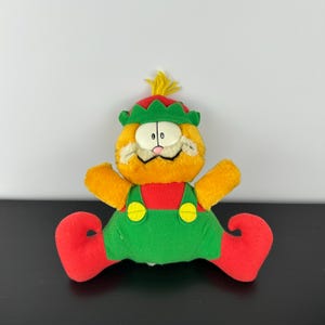 May include: A plush Garfield toy dressed as an elf. The orange cat wears a red and green outfit with a matching hat and elf shoes. The toy has yellow arms and a tuft of yellow hair.