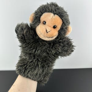 May include: A plush monkey hand puppet with a tan face, ears, and hands, and dark gray fur. The puppet has black eyes and a stitched nose and mouth. The puppet is designed to fit over a hand.