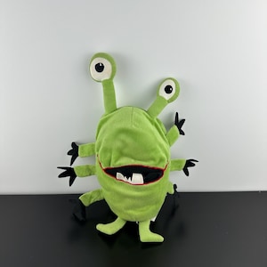 May include: A plush monster toy in a bright lime green color featuring six arms with black hands, two long stalks topped with white and black eyes, and a wide mouth with a red border and white teeth. The creature has a soft, fuzzy texture.
