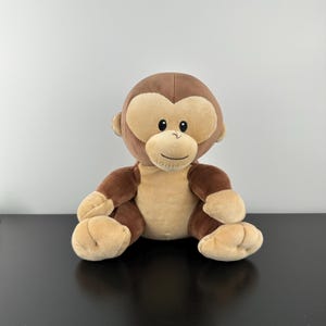 May include: A plush monkey toy with a brown head, arms, and legs, and a tan face, chest, and hands. The monkey has a smiling expression and is sitting upright. The toy is on a black surface against a white background.