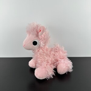 May include: A pink plush toy llama with a fluffy, textured body and a small, rounded head. The llama has a black eye, pink ears, and is sitting on its four legs. The toy is made of soft material.