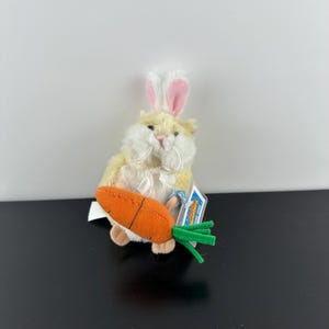 May include: A soft toy bunny with a cream-coloured body, pink and white ears, and a fluffy white face. It holds an orange carrot with green leaves. The toy has a tag attached.