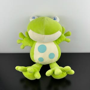 May include: A plush frog toy in shades of green, cream, and blue. The frog has a light green body with a cream-colored belly featuring three blue polka dots. It has large, expressive eyes and outstretched arms and legs.