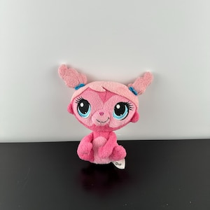 May include: Pink plush toy with blue eyes and pigtails. The toy is sitting with its legs crossed and has a "Shop" tag on its foot.