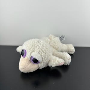 May include: A white plush lamb toy with large purple and black eyes, lying on a black surface. The lamb has a soft, fluffy texture and pink accents on its hooves. A tag is attached to the back of the toy.