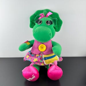 May include: A green plush dinosaur toy wearing a pink vest with a yellow button, a floral skirt, and pink shoes. The dinosaur has large eyes, a pink bow, and a yellow and blue pouch.