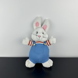 Peut inclure: Une peluche blanche en forme de lapin avec l'intérieur des oreilles roses, des moustaches noires et un nez rose. Le lapin porte une salopette en jean bleu et un haut rayé rouge et blanc. Le jouet a les bras tendus.