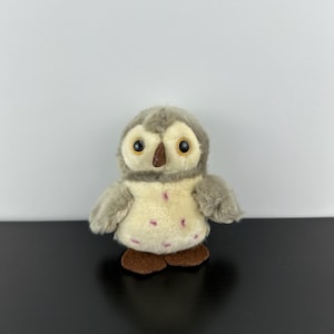 Puede incluir: Un juguete de peluche con forma de búho con cabeza y alas grises, ojos grandes amarillos y pico marrón. El pecho del búho es de color crema con manchas rosas y tiene patas marrones. El juguete está diseñado para parecerse a un bebé búho.