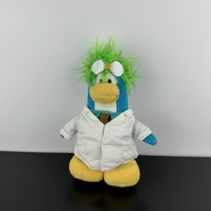 Op de afbeelding: Een pluche pinguïn speelgoed met blauwe en gele kleuren, met een witte labjas, groen haar en een bril. De pinguïn heeft gele voeten en een gele snavel. Het speelgoed staat op een zwart oppervlak tegen een witte achtergrond.