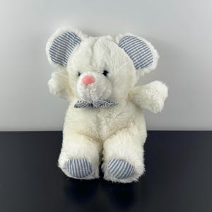 Può includere: Un topolino di peluche bianco con orecchie, piedi e papillon a righe blu. Il topolino ha il naso rosa ed è seduto con un braccio alzato. Il giocattolo è realizzato in materiale morbido e soffice.