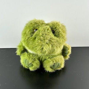 May include: A plush green frog toy with a fuzzy texture. The frog has large, round eyes and a wide, toothy smile.