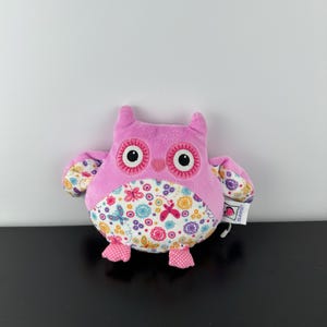Può includere: Un peluche a forma di gufo rosa con grandi occhi neri e bianchi, piccole orecchie e ali decorate. La pancia del gufo presenta un colorato motivo floreale e a farfalle. Il gufo ha piedini rosa a pois bianchi.