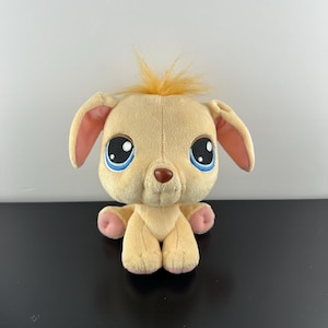 Puede incluir: Un perro de peluche de color beige claro con grandes ojos azules y una nariz marrón. El perro tiene orejas caídas con el interior rosa y un mechón de pelo naranja. Sus patas también son rosas.