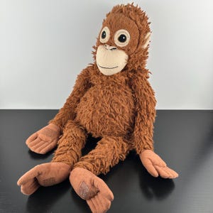 Pode incluir: Um brinquedo de pelúcia de orangotango com pelo castanho felpudo, rosto bege claro e olhos grandes e redondos. O bicho de pelúcia tem braços e pernas longos com mãos e pés planos em castanho claro, com pequenos remendos retangulares nas palmas.