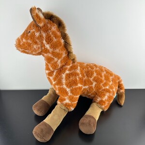 May include: A plush giraffe toy featuring a soft, fuzzy orange and white patterned coat. The stuffed animal has a dark brown mane, tan lower legs, and dark brown hooves. It is depicted in a seated position with its head turned to the left.