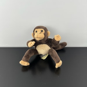 May include: A plush monkey toy with brown fur, tan face, hands, and feet. The stuffed animal has a light tan belly and is posed with its arms and legs spread out. The monkey has a friendly expression and is sitting on a black surface.
