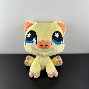 Puede incluir: Un cerdito de peluche con cuerpo amarillo, orejas y hocico naranjas, y ojos grandes con detalles azules y estrellas blancas. El cerdo tiene brazos y piernas pequeños con pies estampados en naranja y amarillo.