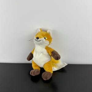 May include: A soft toy fox with a white stomach, brown paws and arms, and a fluffy tail. The fox has a yellow-orange body, white ears, and black whiskers. The toy is on a black surface against a white background.