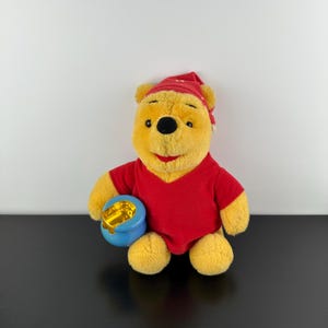 Könnte beinhalten: Eine Plüschpuppe von Winnie Puuh mit rotem Hemd und Nachtmütze, die einen blauen Honigtopf hält. Der Bär hat gelbes Fell, eine schwarze Nase und einen lächelnden Gesichtsausdruck. Die Puppe steht auf einer schwarzen Oberfläche.
