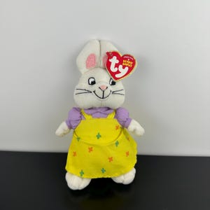 Könnte beinhalten: Ein weißes Plüschhasen-Spielzeug mit langen Ohren und rosafarbenen Innenseiten. Der Hase trägt ein lavendelfarbenes Kurzarmshirt unter einem gelben Trägerkleid mit einem kleinen orangefarbenen und grünen Kreuzmuster. Ein rotes Ty-Herz-Etikett ist befestigt.
