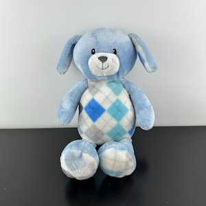 May include: A light blue plush dog with floppy ears and a white and gray argyle pattern on its chest. The dog has a white muzzle, a black nose, and small black eyes. The dog's paws and ears have a matching argyle pattern.