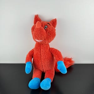 May include: A plush toy fox in bright orange with blue accents on the feet and hands. The fox has a small pink mouth and a red nose. The toy is standing upright against a white and black background.