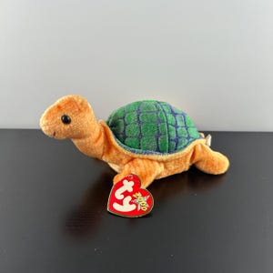 May include: A green and purple turtle beanie baby with a brown shell and orange body. The beanie baby has a red Ty tag with a white star and the text "Beanie Babies" on it.