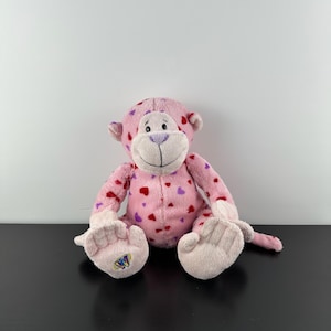 Könnte beinhalten: Ein rosa Plüschaffe mit roten und lila Herzmustern. Der Affe hat ein hellrosa Gesicht, Hände und Füße. Der Affe sitzt auf einer schwarzen Oberfläche. Der Affe hat die Buchstaben "W" und "W" auf seinem linken Fuß gestickt.