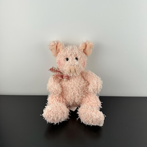 Könnte beinhalten: Ein rosa Plüschschwein mit einem strukturierten, flauschigen Körper. Das Schwein hat kleine schwarze Augen, rosa Ohren und eine rot-weiß karierte Schleife. Das Schwein sitzt aufrecht.