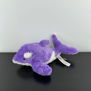 May include: A purple and white plush orca whale toy. The soft toy has a fuzzy texture and a small tag attached. The orca has white patches near the eyes and a white belly. The toy is designed to resemble a killer whale.
