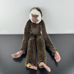 May include: A plush monkey toy with brown fur, a white face, and pink hands and feet. The monkey has long arms and legs, and its eyes are dark brown. The toy is sitting upright.