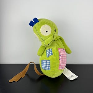 May include: A lime green plush toy with a whimsical design. It features a large, off-white eye, blue accents, and patches of blue gingham and pink gingham fabric. A brown tail extends from the toy.