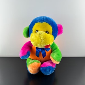 May include: A colourful plush monkey toy. The monkey has a blue head, yellow face, and a red tongue. Its body is a mix of orange, pink, green, and blue. A blue ribbon is tied around its neck.