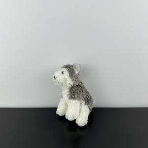 May include: A plush toy dog in a seated position. The dog has a gray and white color scheme, with gray fur on its head and back, and white fur on its chest, legs, and face. The toy is on a black surface.
