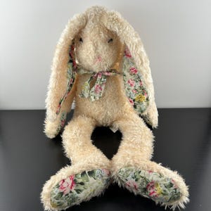 Può includere: Un coniglio di peluche bianco con lunghe orecchie floppy. Le orecchie sono rivestite con un tessuto floreale con fiori rosa, gialli e verdi. Il coniglio ha un nastro floreale intorno al collo e tessuto floreale sul fondo dei piedi.