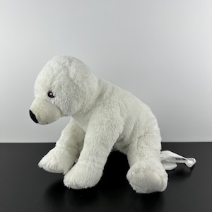 Könnte beinhalten: Ein weißes Plüsch-Eisbär-Spielzeug mit schwarzer Nase und kleinen schwarzen Augen. Das Stofftier befindet sich in einer Krabbelposition auf einer schwarzen Oberfläche. Ein weißes Etikett ist am Spielzeug befestigt.