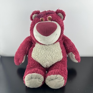 May include: A plush toy bear in shades of pink and cream. The bear has a large, heart-shaped nose, a cream-colored belly, and gray accents on its paws and ears. The bear is smiling and has brown eyes.