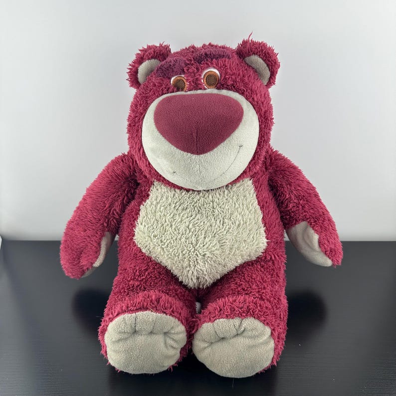 May include: A plush toy bear in shades of pink and cream. The bear has a large, heart-shaped nose, a cream-colored belly, and gray accents on its paws and ears. The bear is smiling and has brown eyes.