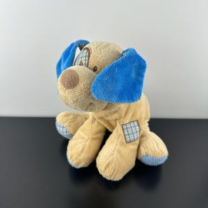 Puede incluir: Un juguete de peluche con forma de perro con pelaje marrón claro, orejas azules y una nariz marrón. El perro tiene un parche a cuadros en la pata y detalles a cuadros en las patas. El juguete está sobre una superficie negra.