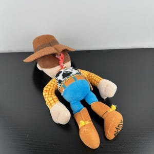 May include: A soft toy of the character Woody from Toy Story. Woody is dressed in a brown cowboy hat, a yellow plaid shirt, blue trousers, and brown boots. The boots have the word "ANDY" written on the sole.