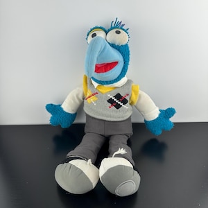 Könnte beinhalten: Ein Plüschtier der Figur Gonzo mit blauem, flauschigem Fell, einer langen, gebogenen Nase und großen weißen Augen. Die Figur trägt eine graue Pullunderweste mit schwarz-gelbem Rautenmuster, weiße Ärmel, eine graue Hose und weiße Schuhe.