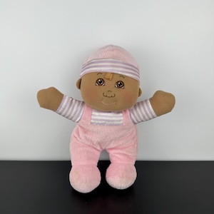 May include: A plush doll with brown skin, brown eyes, and a stitched mouth. The doll wears a pink hat with white stripes, a long-sleeved shirt with pink and white horizontal stripes, and pink overalls. The doll has its arms outstretched.