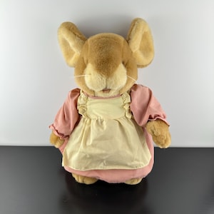 Puede incluir: Un peluche de ratón con grandes orejas y suave pelaje marrón. El ratón lleva un vestido rosa y un delantal amarillo. El juguete está diseñado para parecerse a un personaje de un cuento infantil.