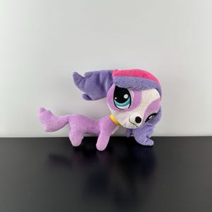 Puede incluir: Peluche de perro morado y blanco con orejas rosas y un collar amarillo. El juguete tiene ojos azules grandes y una nariz blanca.