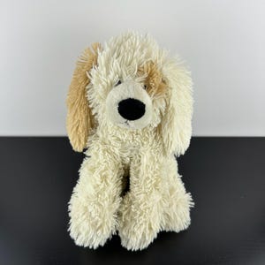 May include: A plush dog toy with cream-colored fur and tan ears. The dog has a black nose and a friendly expression. The toy is sitting upright, showcasing its fluffy texture and soft appearance. A cuddly and comforting children's toy.