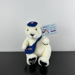 May include: A Hallmark plush polar bear wearing a blue postal worker hat and messenger bag. The bear has black paws and the words "Bear Mail" on the tag. The messenger bag has the words "Postes Canada" printed on it.