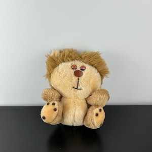 May include: A soft toy lion with a tan body, brown mane, and brown button eyes. The lion has small, rounded paws with brown spots. The toy is sitting on a black surface against a white background.